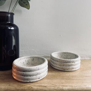 May include: Two white ceramic bowls with a speckled finish and raised edges. The bowls are on a light wood surface.