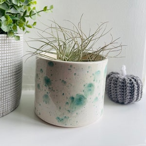 May include: A white ceramic planter with green splatter paint design, featuring a small air plant inside. The planter is sitting on a white surface next to a grey crocheted pumpkin.