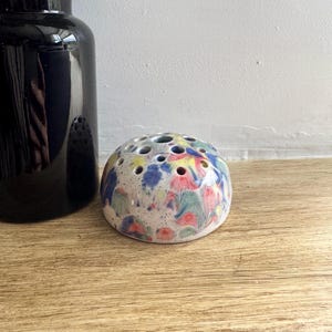 May include: A ceramic flower frog with a speckled, colourful design. The frog is round with multiple holes for arranging flowers. The colours include blue, red, yellow, and green. A black vase is in the background.