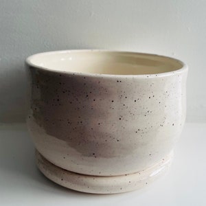 May include: A white ceramic planter with a speckled finish and a matching saucer. The planter has a rounded shape and a smooth, glossy surface.