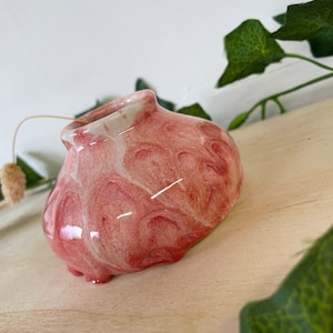 May include: A small, pink and white ceramic vase with a marbled glaze. The vase has a rounded, slightly irregular shape and a wide opening.