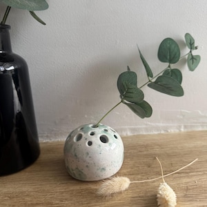 May include: A small, round, ceramic bud vase with a speckled white and pale green finish. The vase has several holes on top for holding stems. A sprig of green eucalyptus and dried flowers are arranged in the vase. A black vase is in the background.