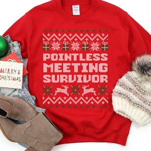 May include: A red sweatshirt with a festive pattern and the words "POINTLESS MEETING SURVIVOR" in white. The design includes snowflakes, gift boxes, and reindeer. A green Christmas ornament, a "Merry Christmas" card, and a knit hat are also in the image.