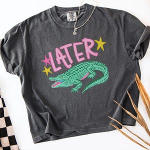 Peut inclure: T-shirt gris fonc&eacute; avec un motif d'alligator vert et le mot "LATER" en rose, avec des &eacute;toiles jaunes. Le t-shirt a un aspect vintage et us&eacute;. Un collier argent&eacute; est drap&eacute; sur le t-shirt.