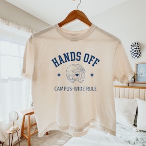 May include: Cream-colored t-shirt with the text "HANDS OFF" above a helmet graphic and the words "CAMPUS-WIDE RULE" below. The text and graphic are in navy blue. The shirt is hanging on a wooden hanger.