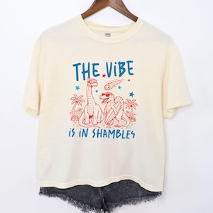 May include: Cream-coloured t-shirt featuring a blue and red graphic of two dinosaurs, palm trees, and a shooting star. The text "THE VIBE is in SHAMBLES" is printed above the dinosaurs. The shirt is hanging on a wooden hanger.