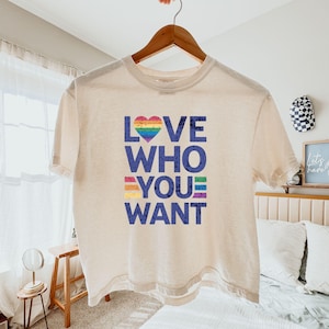 Puede incluir: Una camiseta color crema con el texto "LOVE WHO YOU WANT" en letras azules. La palabra "LOVE" presenta un corazón arcoíris, y las palabras "YOU WANT" tienen rayas arcoíris. La camiseta está colgada en una percha de madera.