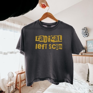 Puede incluir: Una camiseta de manga corta de color gris carbón con el texto "radical left scum" impreso en amarillo con una tipografía estilo nota de rescate en el pecho. La camiseta tiene cuello redondo y se muestra en una percha de madera.
