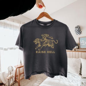 May include: Dark gray t-shirt with a gold graphic of a person riding a tiger, with the words "RAISE HELL" printed below. The shirt is hanging on a wooden hanger.