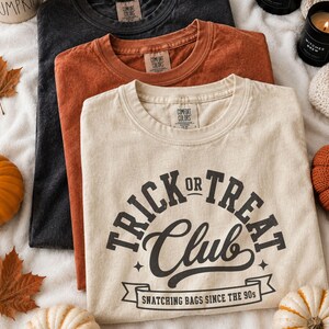 May include: Three crew neck t-shirts in black, burnt orange, and cream. The cream shirt features black text reading "TRICK OR TREAT CLUB" and "SNATCHING BAGS SINCE THE 90S" with star accents and a banner graphic. Each shirt has a Comfort Colors label.