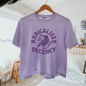 May include: A light purple t-shirt with the words "RADICALIZED BY BASIC DECENCY" encircling a raven holding a branch. The shirt is hanging on a wooden hanger.