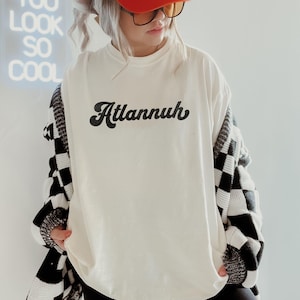 May include: Cream-colored t-shirt with the word "Atlannuh" in black script. The person is wearing an orange cap, sunglasses, and a black and white checkered cardigan. A neon sign in the background reads "You Look So Cool."