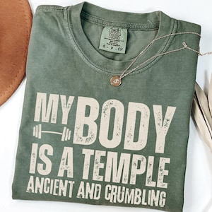 May include: An olive green t-shirt with the text "MY BODY IS A TEMPLE ANCIENT AND CRUMBLING" in white block letters. A barbell graphic is above the words "IS A". A gold necklace with a pendant is on the shirt. A brown hat is in the upper left corner.