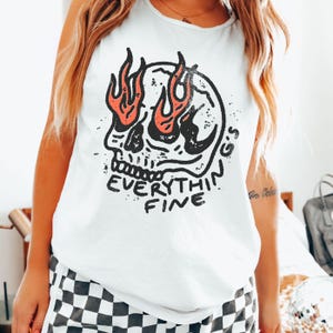 Op de afbeelding: Een wit mouwloos hemd met een zwarte afbeelding van een schedel met oranje vlammen uit de oogkassen. Onder de schedel staat de tekst "EVERYTHING'S FINE" gedrukt in een verweerd zwart lettertype. Het shirt heeft een ronde halslijn.