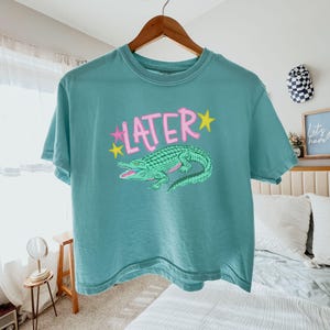 Peut inclure: T-shirt de couleur sarcelle avec le mot "LATER" en rose et des &eacute;toiles jaunes. Un dessin d'alligator vert est en dessous. Le t-shirt est suspendu &agrave; un cintre en bois. V&ecirc;tement d&eacute;contract&eacute;.
