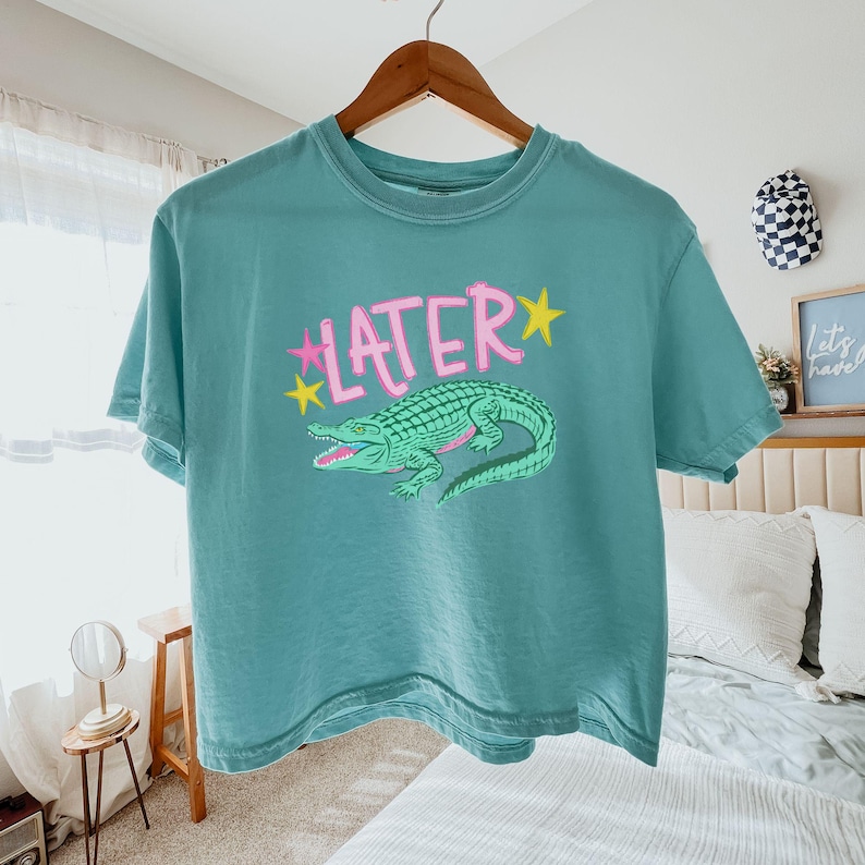 Peut inclure: T-shirt de couleur sarcelle avec le mot "LATER" en rose et des &eacute;toiles jaunes. Un dessin d'alligator vert est en dessous. Le t-shirt est suspendu &agrave; un cintre en bois. V&ecirc;tement d&eacute;contract&eacute;.