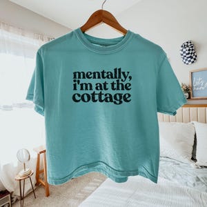 Op de afbeelding: Een turquoise t-shirt met de tekst "mentally, I'm at the cottage" in het zwart. Het shirt hangt aan een houten hanger. Het t-shirt is een casual kledingstuk.