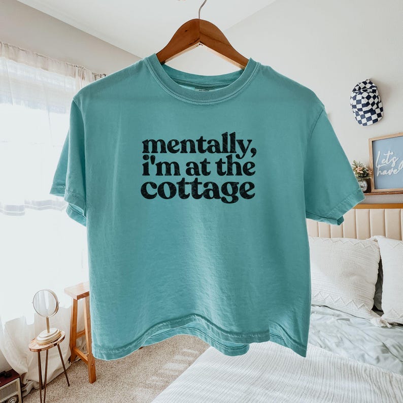 Op de afbeelding: Een turquoise t-shirt met de tekst "mentally, I'm at the cottage" in het zwart. Het shirt hangt aan een houten hanger. Het t-shirt is een casual kledingstuk.