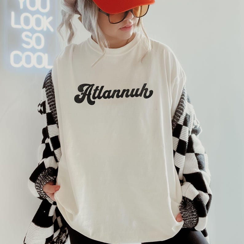 May include: Cream-colored t-shirt with the word "Atlannuh" in black script. The person is wearing an orange cap, sunglasses, and a black and white checkered cardigan. A neon sign in the background reads "You Look So Cool."