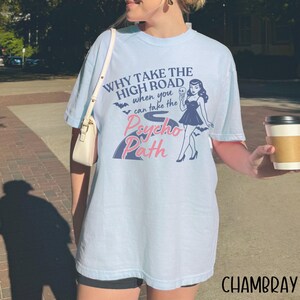 May include: Light blue t-shirt with the text "WHY TAKE THE HIGH ROAD when you can take the Psycho Path" and a graphic of a woman in a black dress. The word "CHAMBRAY" is printed in the bottom right corner.