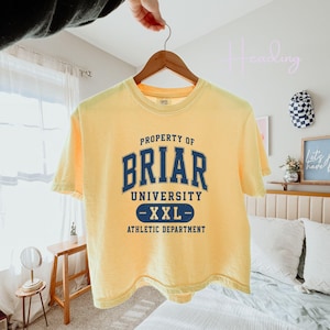 May include: A yellow t-shirt with the text "PROPERTY OF BRIAR UNIVERSITY - XXL - ATHLETIC DEPARTMENT" in navy blue. The shirt is hanging on a wooden hanger.