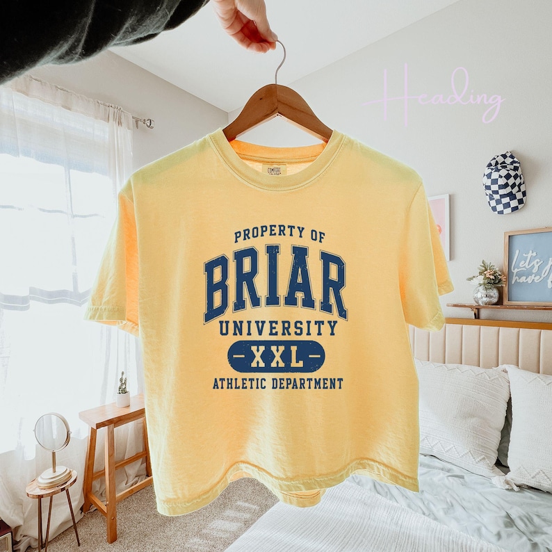 May include: A yellow t-shirt with the text "PROPERTY OF BRIAR UNIVERSITY - XXL - ATHLETIC DEPARTMENT" in navy blue. The shirt is hanging on a wooden hanger.