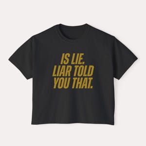 K&ouml;nnte beinhalten: Schwarzes T-Shirt mit dem goldenen Text "IS LIE. LIAR TOLD YOU THAT.". Das T-Shirt mit Rundhalsausschnitt ist ein schlichtes Design, ideal f&uuml;r den Alltag. Der Text ist mittig auf der Vorderseite platziert.