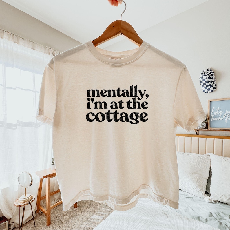 Op de afbeelding: Cr&egrave;mekleurig T-shirt met de zwarte tekst "mentally, I'm at the cottage". Het T-shirt hangt aan een houten kleerhanger.