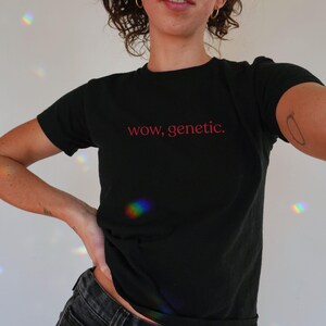 May include: A black short-sleeved t-shirt featuring the text "wow, genetic." printed in a red, lowercase sans-serif font across the center of the chest. The garment has a classic crew neckline and a standard fit, made of a solid black fabric.