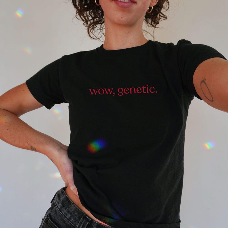 May include: A black short-sleeved t-shirt featuring the text "wow, genetic." printed in a red, lowercase sans-serif font across the center of the chest. The garment has a classic crew neckline and a standard fit, made of a solid black fabric.
