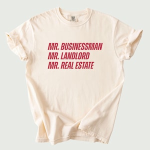 May include: Cream-colored t-shirt with the phrases "MR. BUSINESSMAN", "MR. LANDLORD", and "MR. REAL ESTATE" printed in red. The shirt has short sleeves and a crew neck. The fabric appears soft and comfortable.