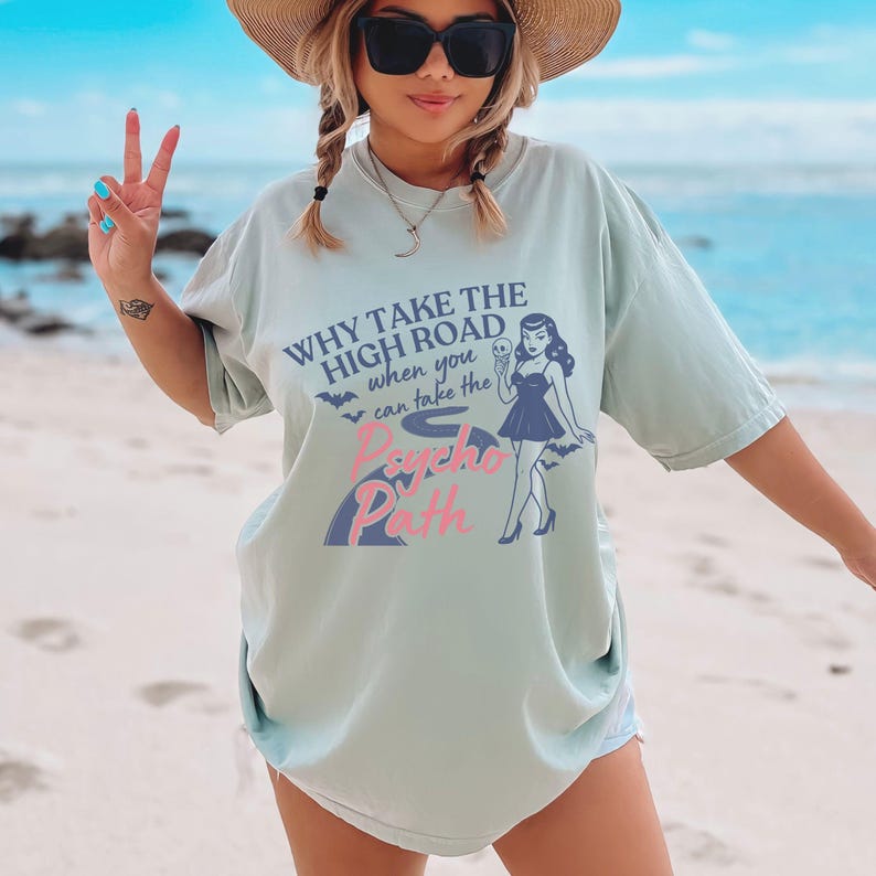 May include: A light green t-shirt with the text "WHY TAKE THE HIGH ROAD when you can take the Psycho Path." The shirt features a retro illustration of a woman holding an ice cream cone. The shirt is a relaxed fit.