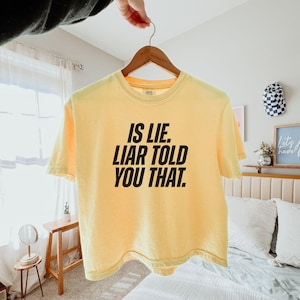 K&ouml;nnte beinhalten: Ein hellgelbes T-Shirt mit dem Text "IS LIE. LIAR TOLD YOU THAT." in fetten schwarzen Buchstaben. Das T-Shirt h&auml;ngt an einem Holzb&uuml;gel.