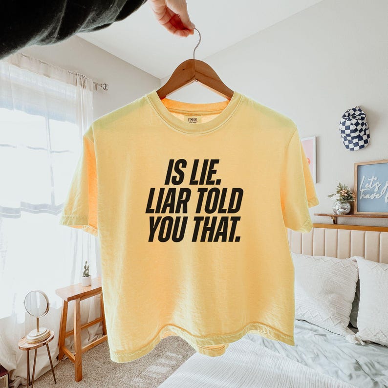 K&ouml;nnte beinhalten: Ein hellgelbes T-Shirt mit dem Text "IS LIE. LIAR TOLD YOU THAT." in fetten schwarzen Buchstaben. Das T-Shirt h&auml;ngt an einem Holzb&uuml;gel.