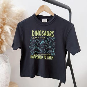 May include: A black t-shirt with the text "DINOSAURS didn't read and we know what HAPPENED TO THEM" in green and blue. The shirt features dinosaur illustrations and book graphics. The t-shirt is hanging on a wooden hanger.