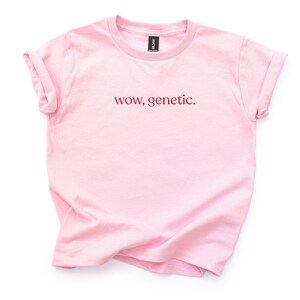 May include: A light pink short-sleeved t-shirt with a crew neckline and rolled sleeves. The center of the chest features the text "wow, genetic." printed in a dark red, lowercase serif font. The garment is displayed flat against a plain background.