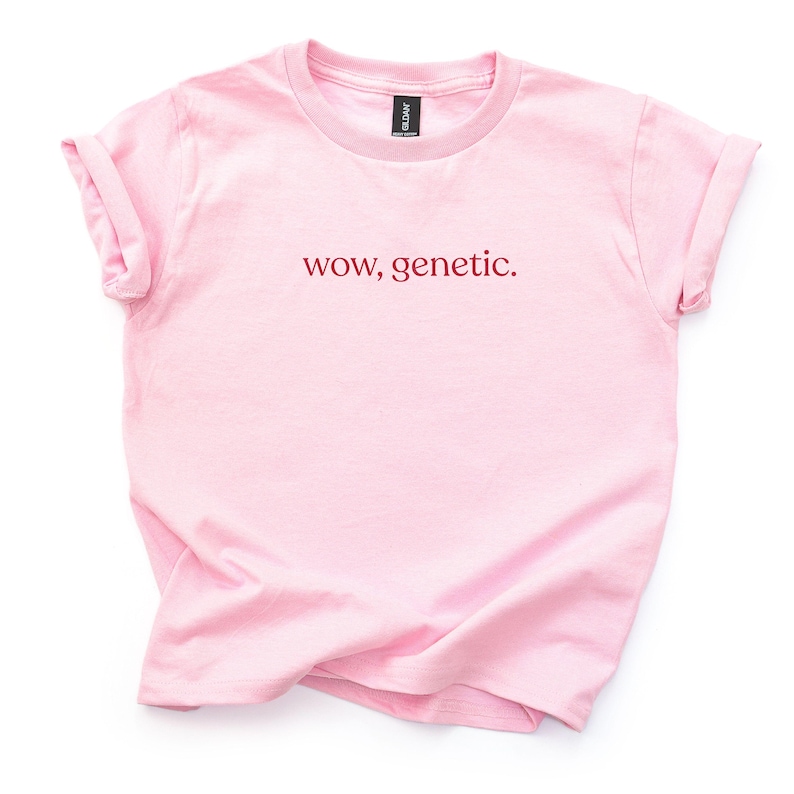 May include: A light pink short-sleeved t-shirt with a crew neckline and rolled sleeves. The center of the chest features the text "wow, genetic." printed in a dark red, lowercase serif font. The garment is displayed flat against a plain background.