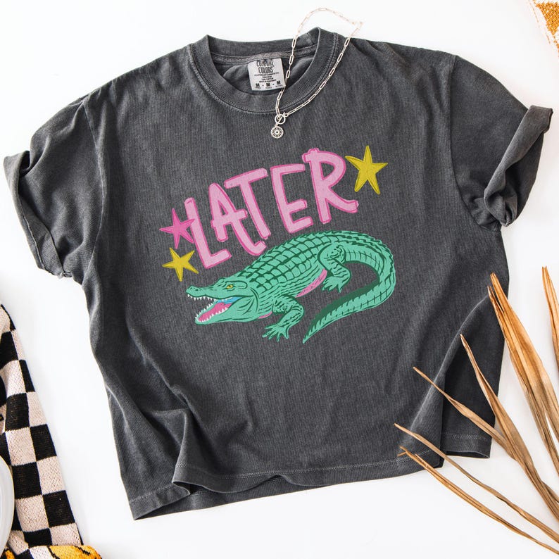 Peut inclure: T-shirt gris fonc&eacute; avec un motif d'alligator vert et le mot "LATER" en rose, avec des &eacute;toiles jaunes. Le t-shirt a un aspect vintage et us&eacute;. Un collier argent&eacute; est drap&eacute; sur le t-shirt.
