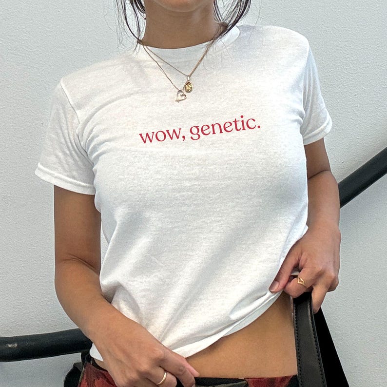 May include: A white short-sleeved t-shirt featuring the text "wow, genetic." printed in a red serif font across the chest. The garment has a crew neckline and is worn with two gold-toned necklaces, one with a heart charm and one with a small pendant.