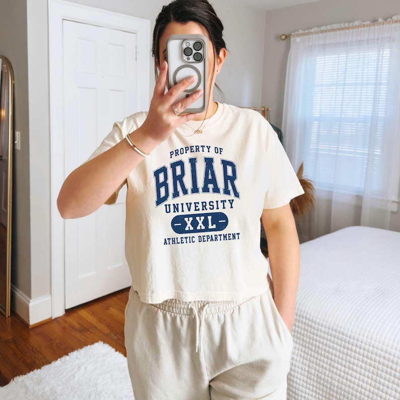 May include: Cream-colored cropped t-shirt with the text "PROPERTY OF BRIAR UNIVERSITY - XXL - ATHLETIC DEPARTMENT" in navy blue. The shirt is paired with light beige sweatpants. The image is taken indoors.