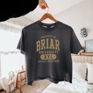 May include: A dark gray t-shirt with the text "PROPERTY OF BRIAR UNIVERSITY - XXL - ATHLETIC DEPARTMENT" in gold lettering. The shirt is hanging on a wooden hanger.