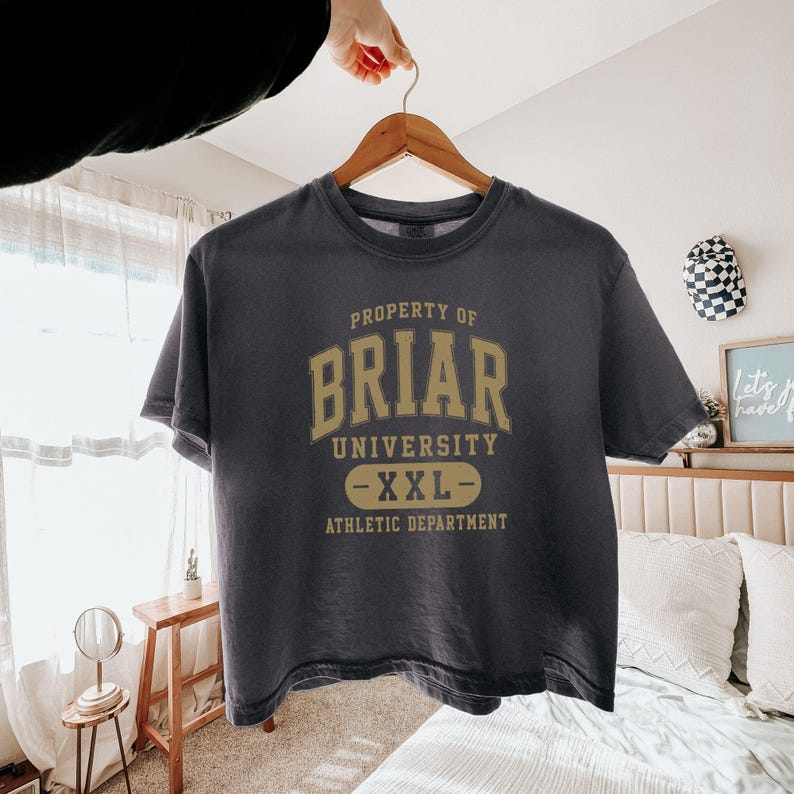 May include: A dark gray t-shirt with the text "PROPERTY OF BRIAR UNIVERSITY - XXL - ATHLETIC DEPARTMENT" in gold lettering. The shirt is hanging on a wooden hanger.