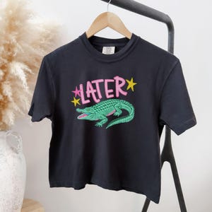 Peut inclure: T-shirt noir avec un motif d'alligator vert et le mot "LATER" en rose, avec des &eacute;toiles jaunes. Le t-shirt est suspendu &agrave; un cintre en bois. Le design est de style d&eacute;contract&eacute; et ludique.