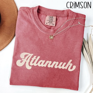 May include: Crimson-colored t-shirt with the word "Atlannuh" in a retro font. A silver necklace with a pendant is draped on the shirt. The shirt is made of Comfort Colors heavyweight garment dye cotton. A brown hat is visible in the background.