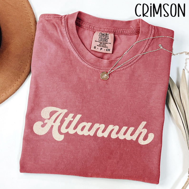 May include: Crimson-colored t-shirt with the word "Atlannuh" in a retro font. A silver necklace with a pendant is draped on the shirt. The shirt is made of Comfort Colors heavyweight garment dye cotton. A brown hat is visible in the background.
