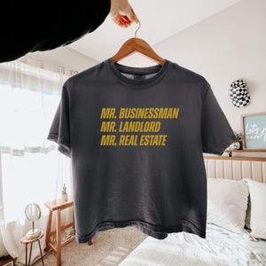 May include: Dark gray t-shirt with the phrases "MR. BUSINESSMAN", "MR. LANDLORD", and "MR. REAL ESTATE" printed in gold. The shirt is hanging on a wooden hanger.