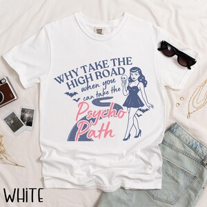 May include: White t-shirt with a retro design featuring a woman holding a skull and the text "WHY TAKE THE HIGH ROAD when you can take the Psycho Path." The design includes bats and a road graphic. Sunglasses and jeans are in the background.