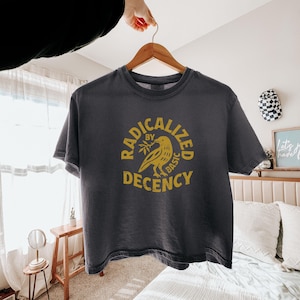 Peut inclure: T-shirt gris fonc&eacute; avec un motif jaune repr&eacute;sentant un oiseau tenant une branche d'olivier et les mots "RADICALIZED BY BASIC DECENCY". Le t-shirt est suspendu &agrave; un cintre en bois.