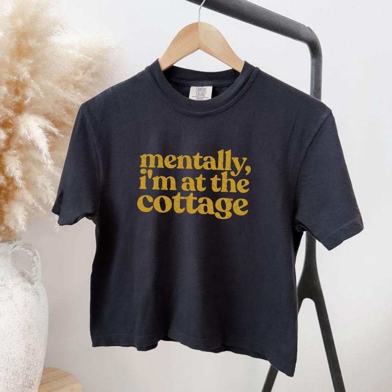 Op de afbeelding: Een zwart T-shirt met de tekst "mentally, I'm at the cottage" in gouden letters. Het shirt hangt aan een houten hanger. Het T-shirt is een cropped model. De achtergrond is neutraal.