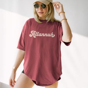 May include: A maroon t-shirt with the word "Atlannuh" in a retro font. The shirt is short-sleeved and has a relaxed fit. The person in the image is wearing a beige baseball cap and sunglasses.